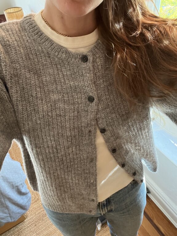 A person wearing a grey knit cardigan over a white shirt and blue jeans, standing indoors near a window.