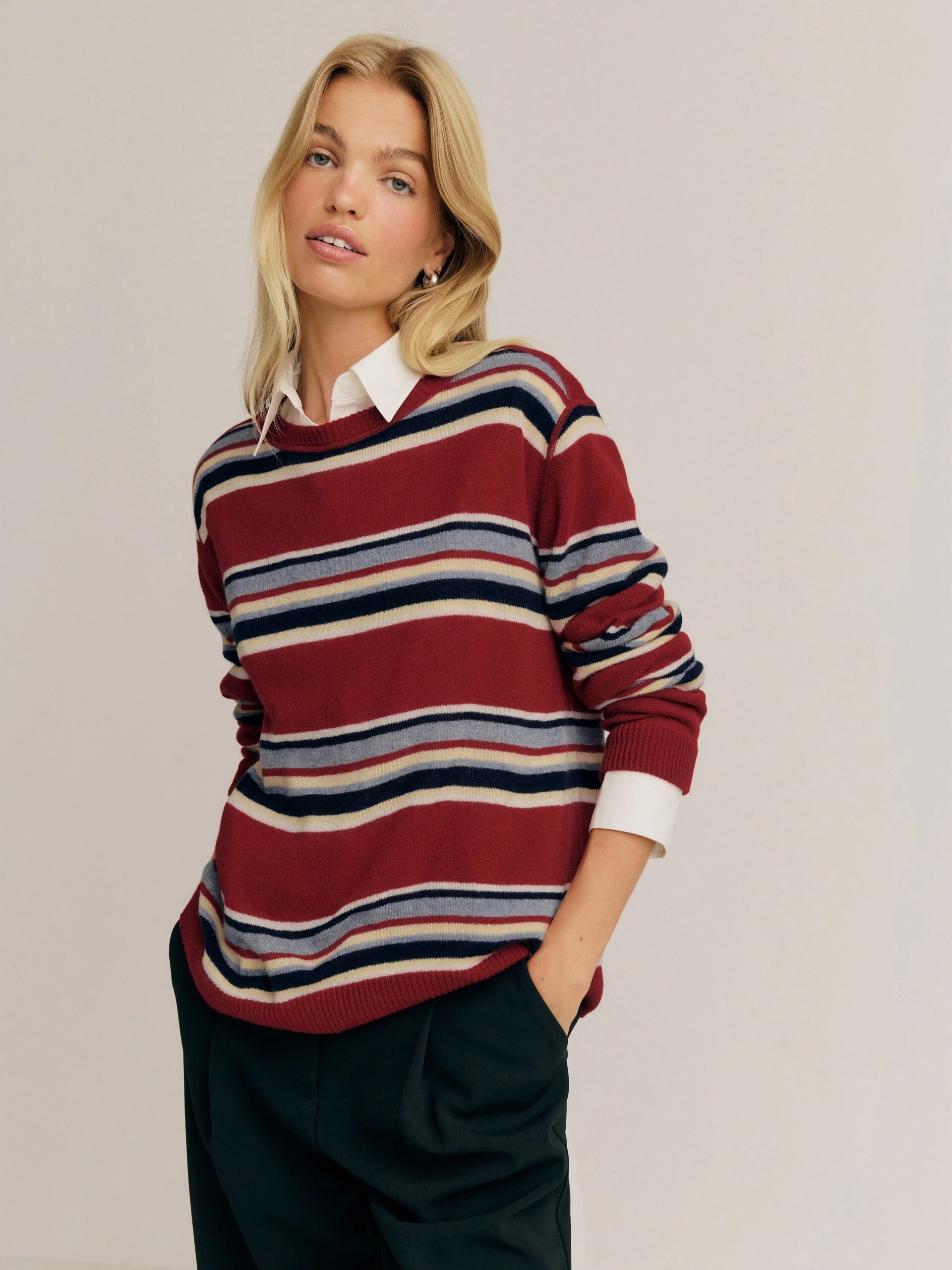A woman stands against a plain background, wearing a red striped sweater over a white collared shirt with dark pants, with one hand in her pocket.