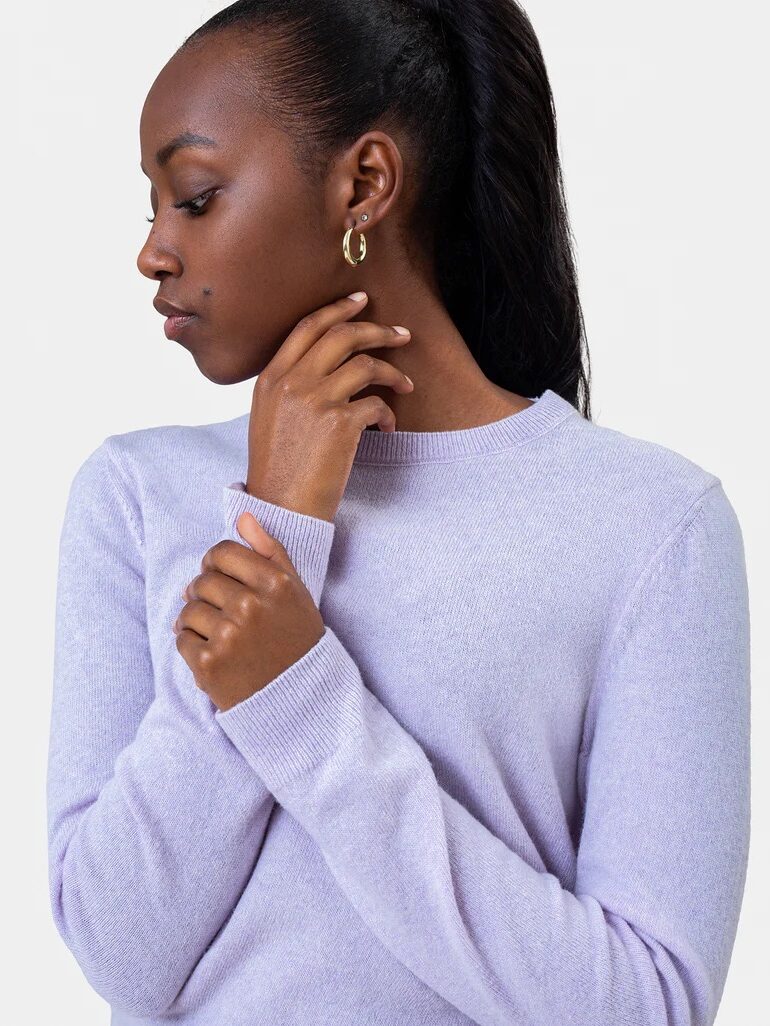 A woman wearing a light purple long-sleeve top looks to her left with one hand touching her neck and the other crossing her chest.