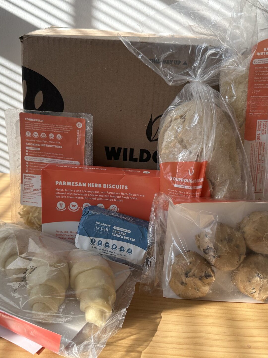 A meal kit delivery box and its contents, including packaged ingredients, recipe cards, biscuits, dough, and other food items, arranged on a wooden table.